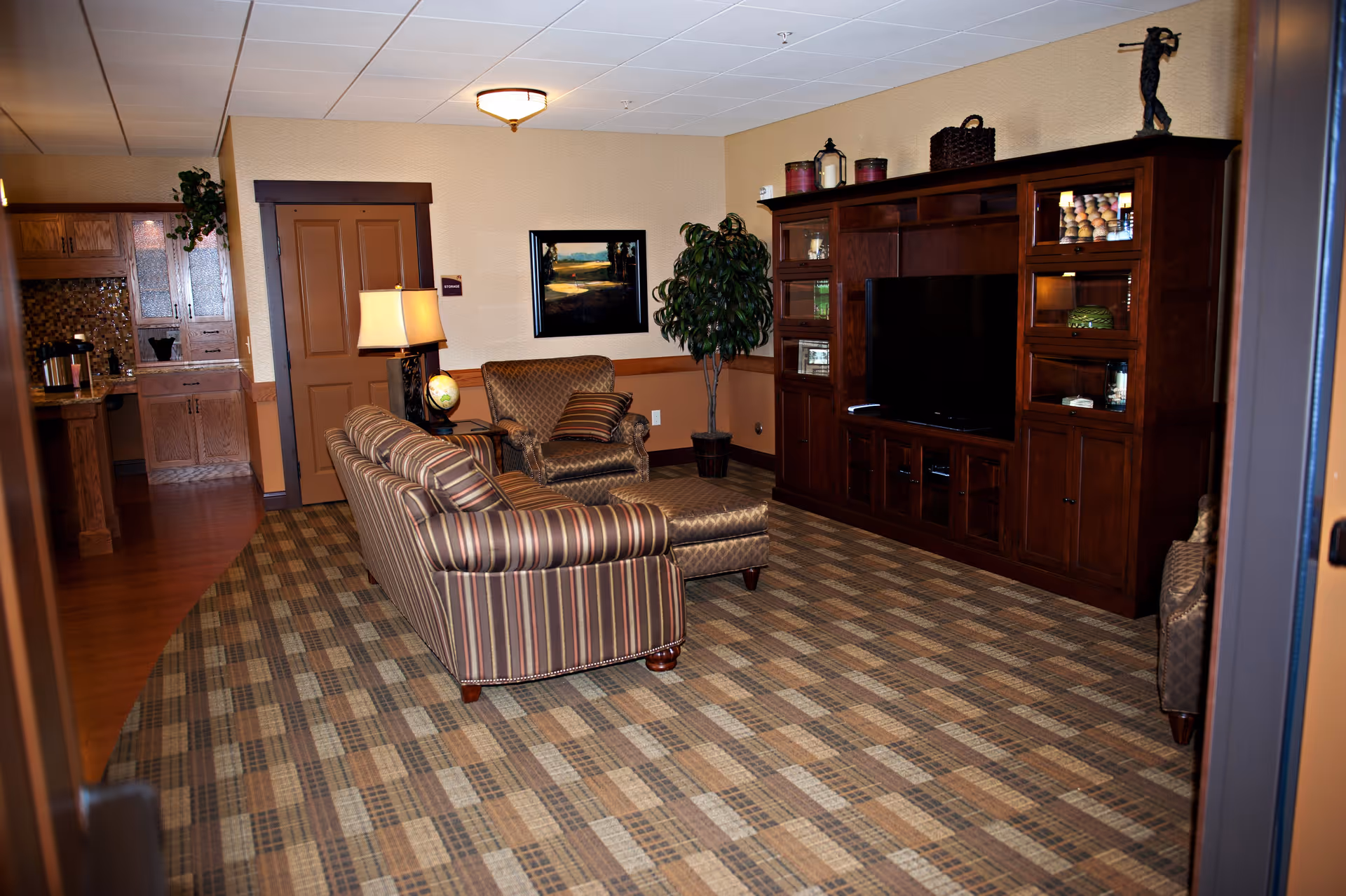 A cozy living room in an assisted living facility featuring a striped sofa, an armchair with a matching ottoman, a wooden entertainment center with a flat-screen TV, decorative items, a potted plant, a table lamp, and a framed landscape painting on the wall. The room has a patterned carpet and warm-toned walls.
