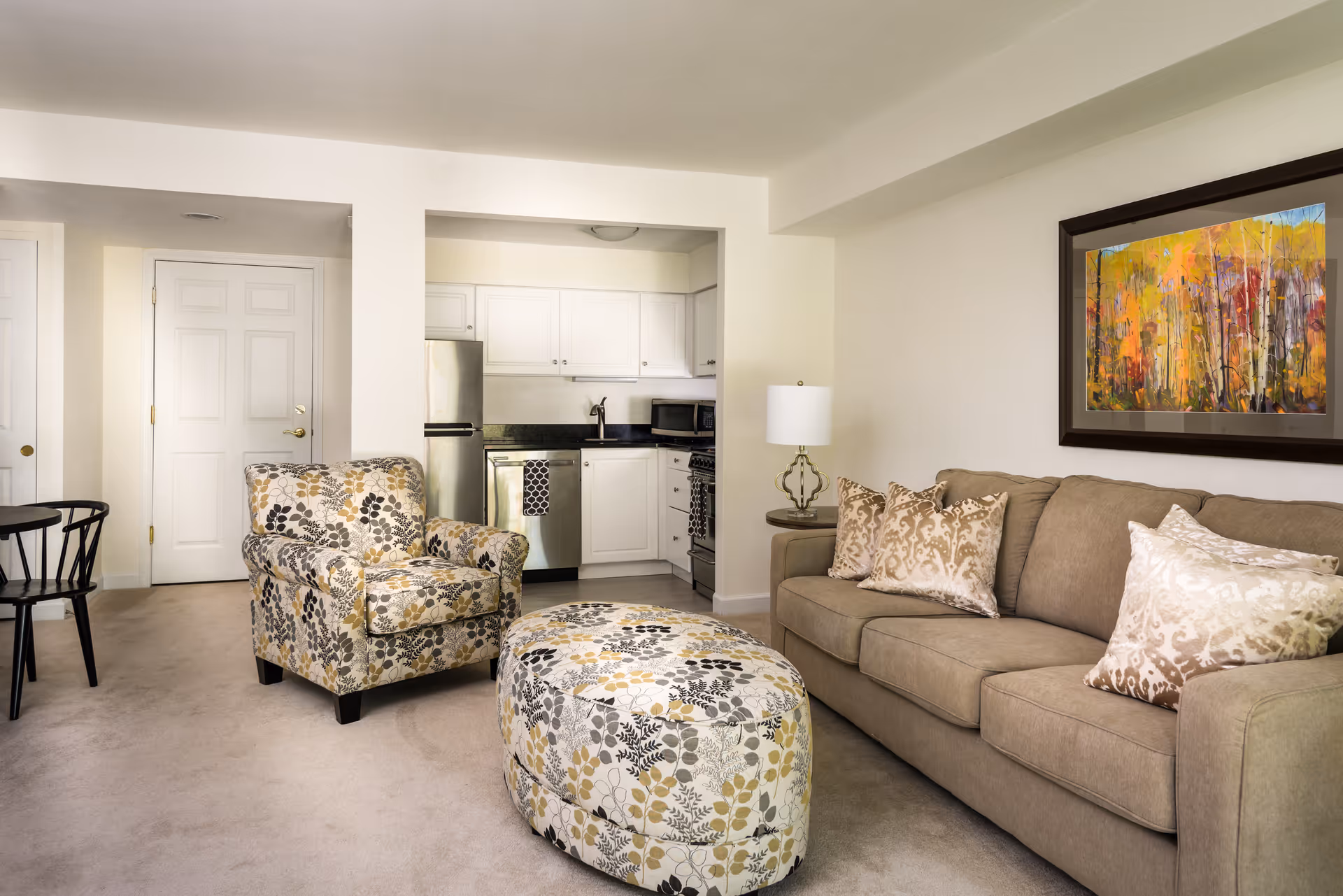 A cozy living room area with a beige sofa adorned with decorative pillows, a matching floral-patterned armchair, and a round floral-patterned ottoman. The room features a small kitchen with white cabinets, a stainless steel refrigerator, dishwasher, and stove. A colorful abstract painting hangs on the wall above the sofa, and a table lamp sits on a side table next to the sofa.