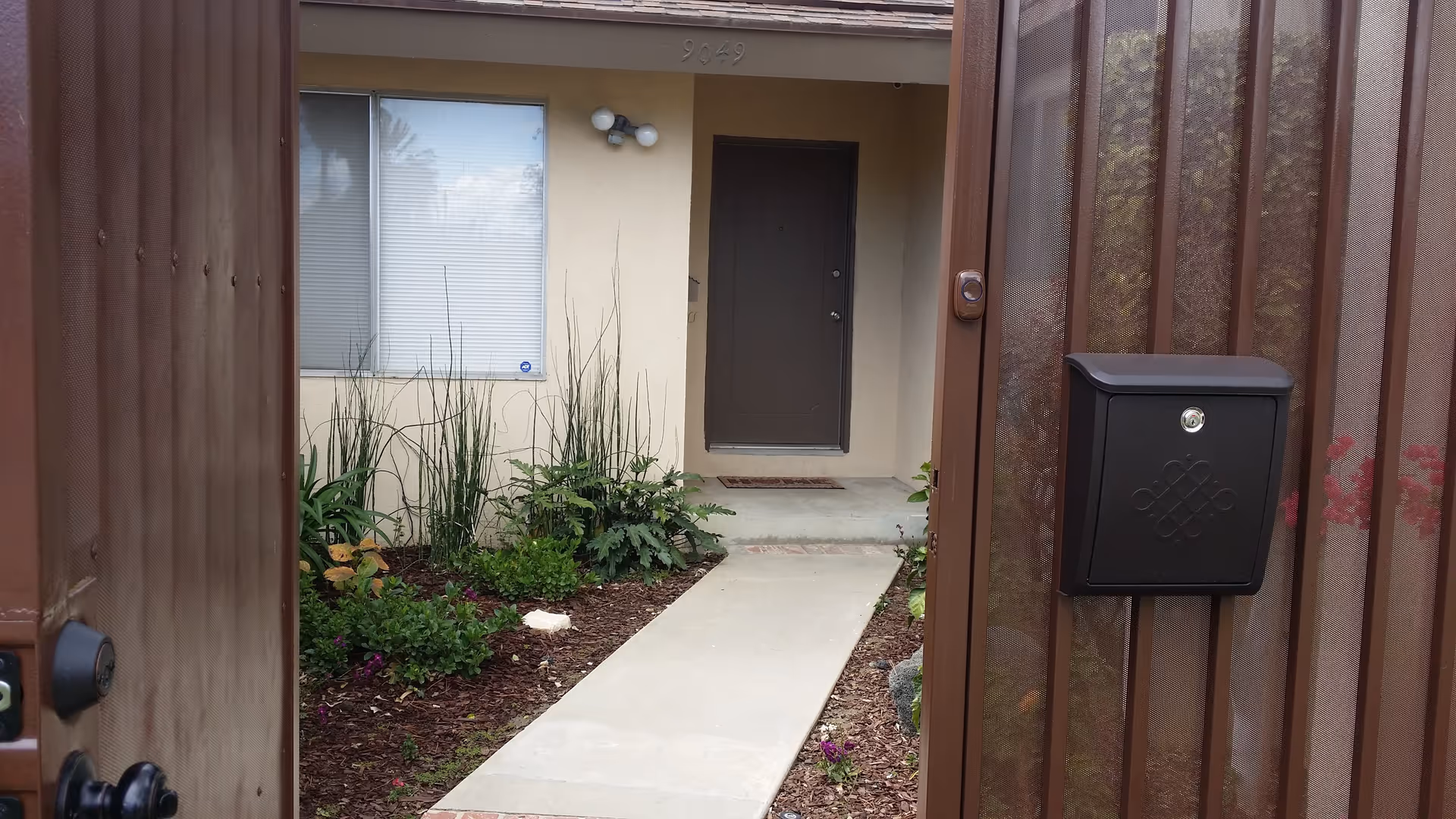 View through an open brown metal gate showing a concrete pathway leading to a dark brown front door of a beige building. There are plants and mulch on either side of the pathway, a window with closed blinds to the left of the door, a black mailbox attached to the gate, and the number 9649 above the door.