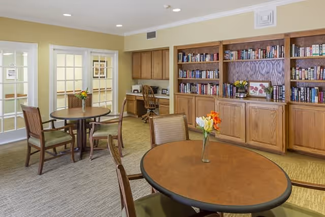 A cozy interior room with round wooden tables and chairs, each table adorned with a small vase of flowers. There is a large wooden bookshelf filled with books along one wall, and a small desk with a chair in the corner. The room has beige walls and carpeted flooring, with glass-paneled double doors leading to another area.