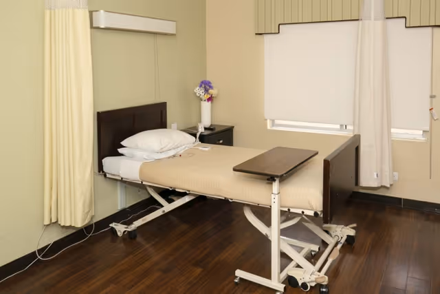 Rehabilitation room with an adjustable hospital bed, overbed table, nightstand with flowers, and closed window shades.