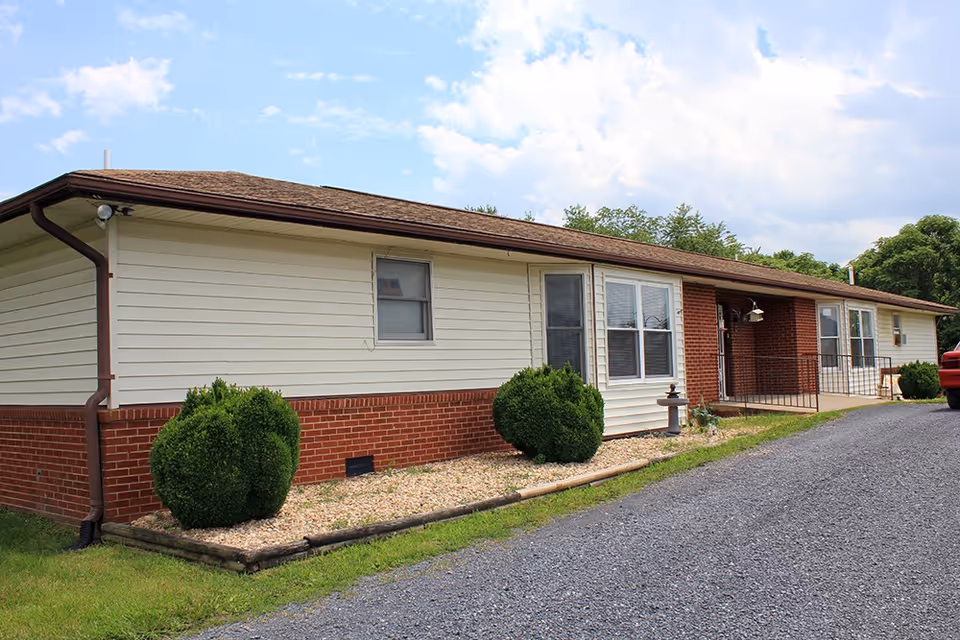 Single-story brick and siding building with shrubs and a gravel driveway under a partly cloudy sky.