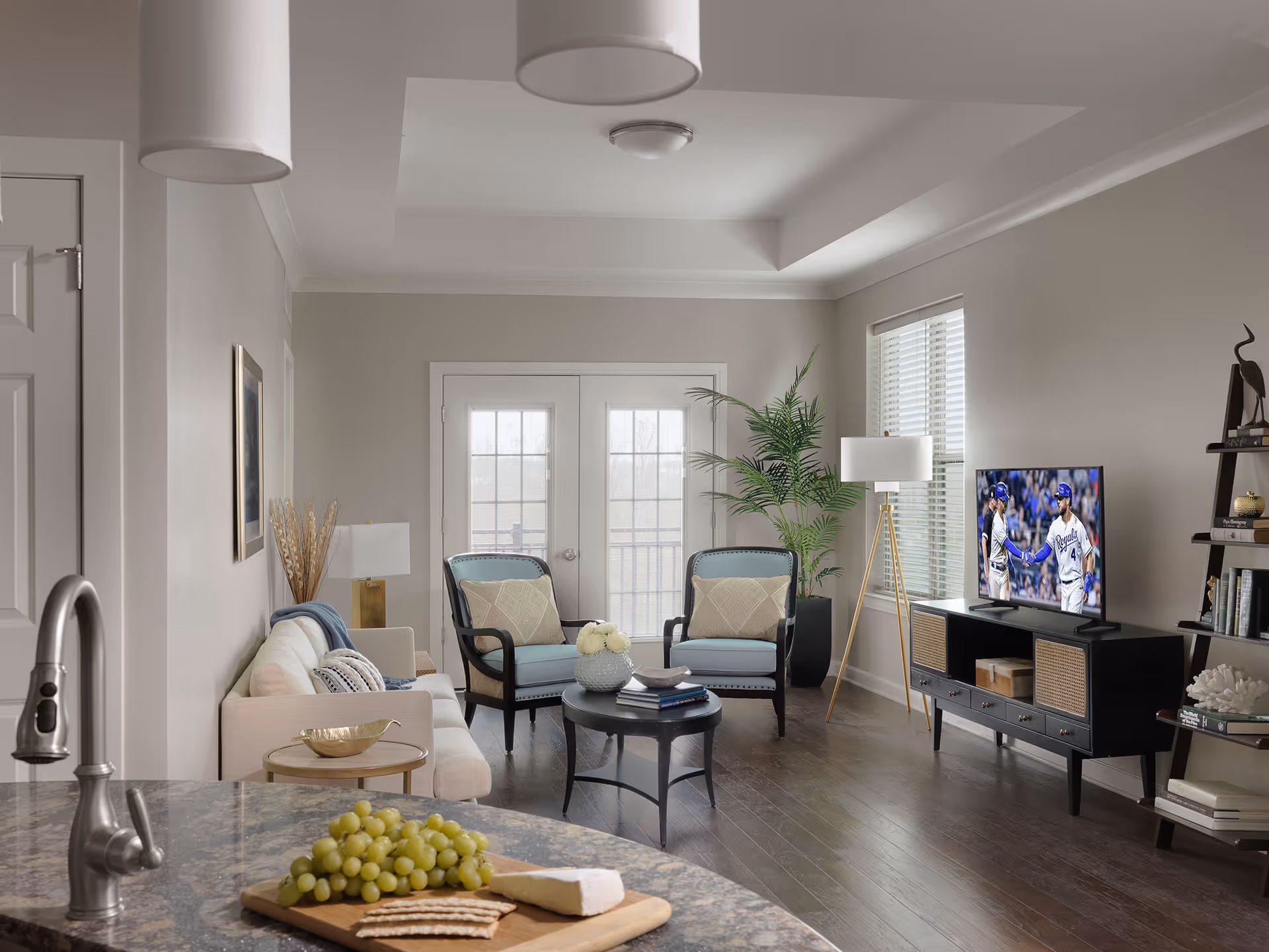 Bright modern living room with a sofa, two armchairs, coffee table, TV on a media console, and a kitchen island with grapes and cheese in the foreground.