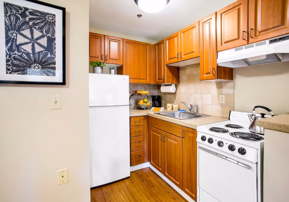 A compact kitchen with wooden cabinets, a white refrigerator, an electric stove with four burners, a stainless steel sink, and a countertop with a coffee maker, a fruit basket, and two mugs. There is a framed black and white abstract artwork on the wall to the left.