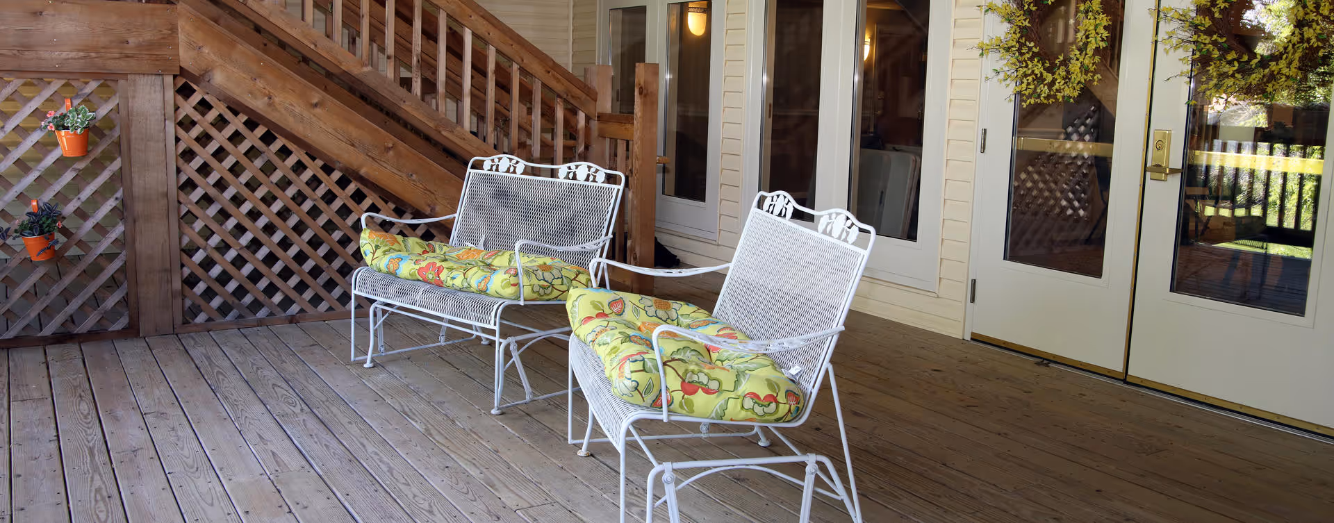 Outdoor wooden deck area with two white metal benches, each with a green floral cushion. There are wooden stairs and lattice railing on the left side, and double glass doors with a wreath on the right side.