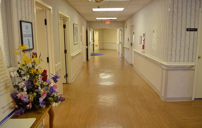 Well-lit interior hallway in a senior living facility with wood-look floors, handrails and doors along the sides and a floral arrangement on a small table.