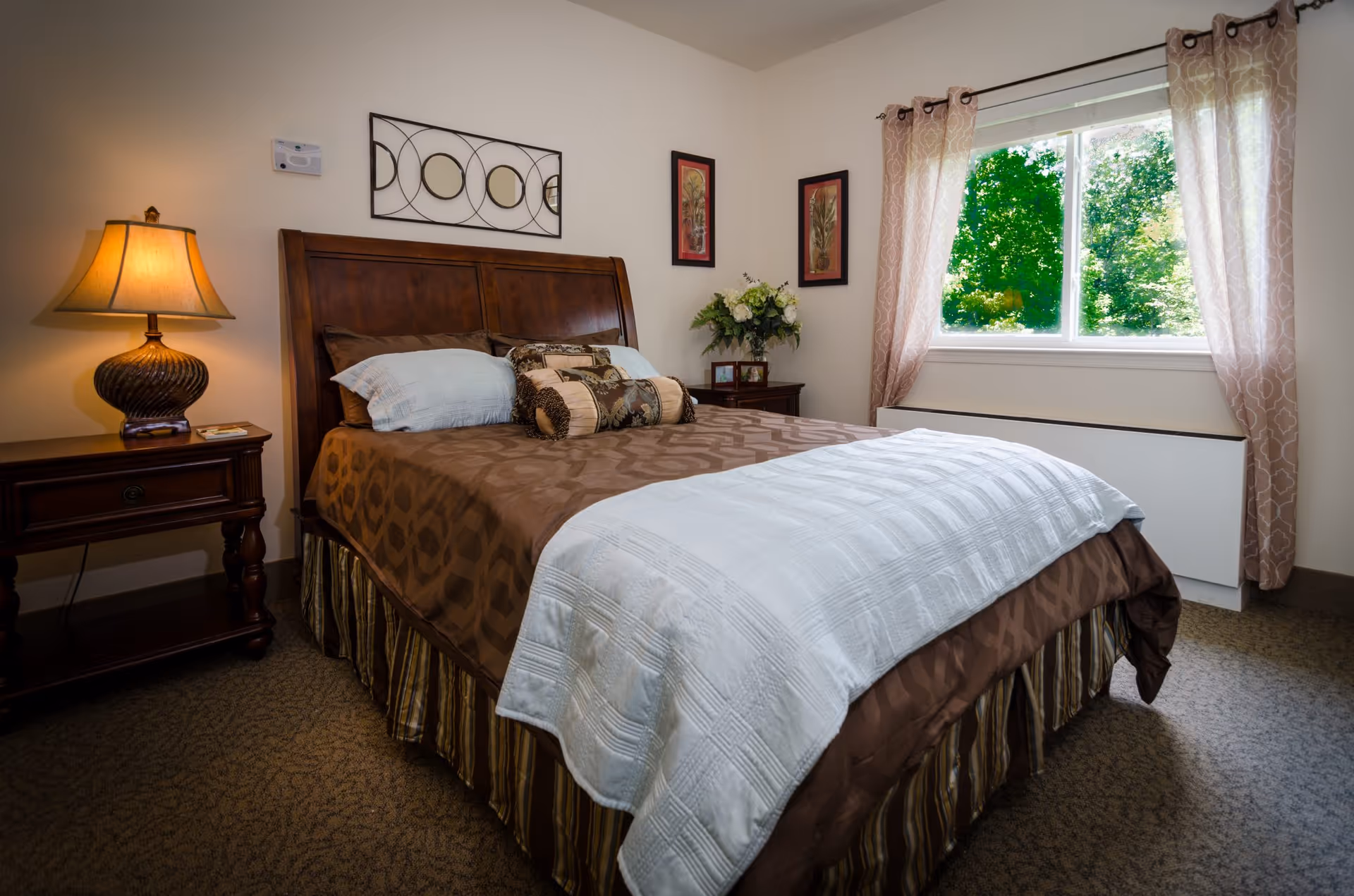 Well-lit bedroom with a made bed featuring a wooden headboard, nightstand with a lamp, window with curtains and a view of trees.