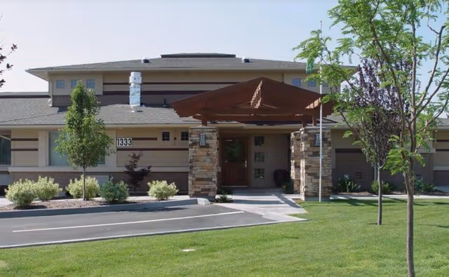 Front entrance of a single-story building with a covered wooden porte-cochere, stone columns, driveway and landscaped lawn.