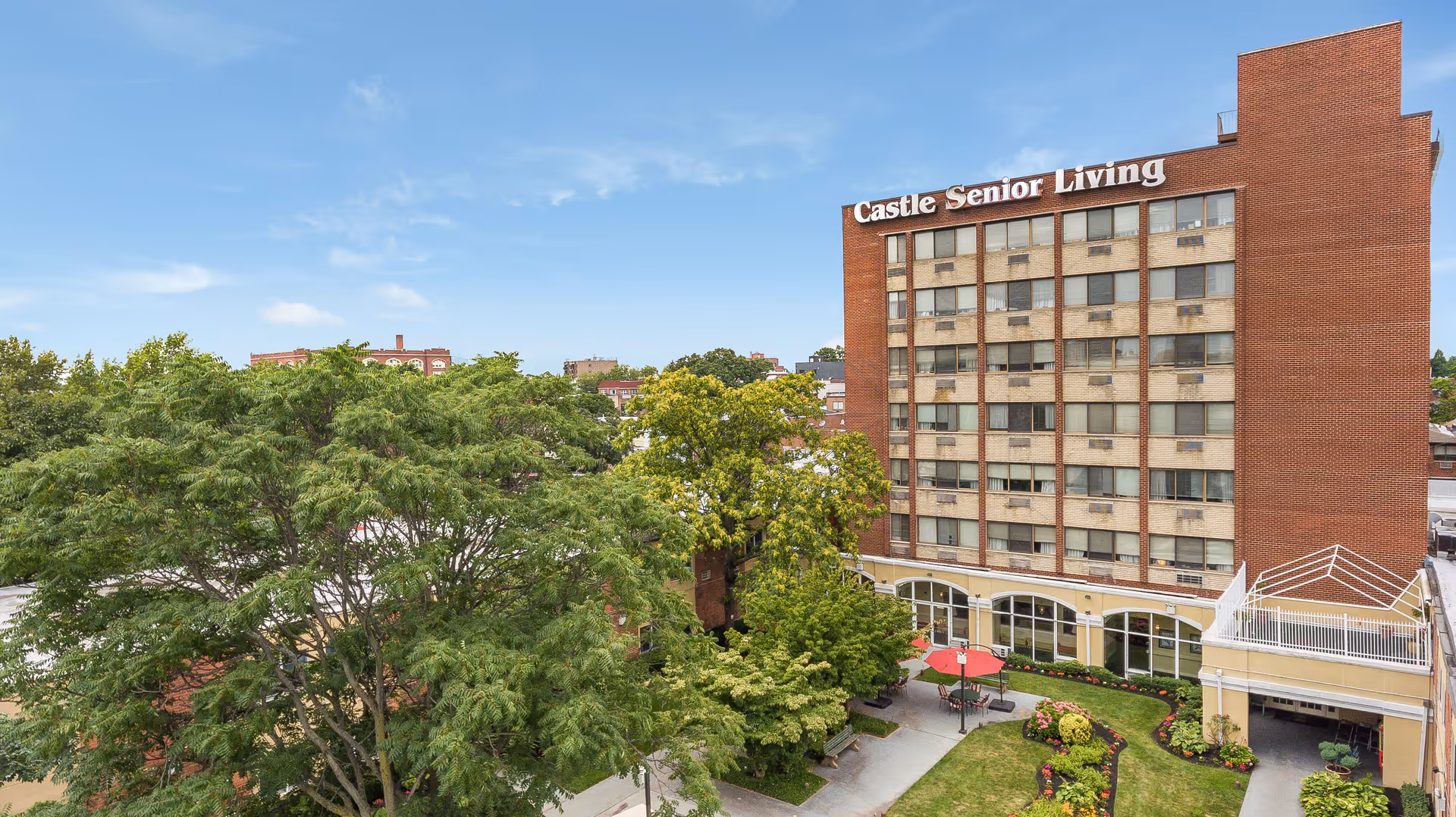 Multi-story red brick building labeled 'Castle Senior Living' with a landscaped courtyard, patio umbrellas, and surrounding trees.
