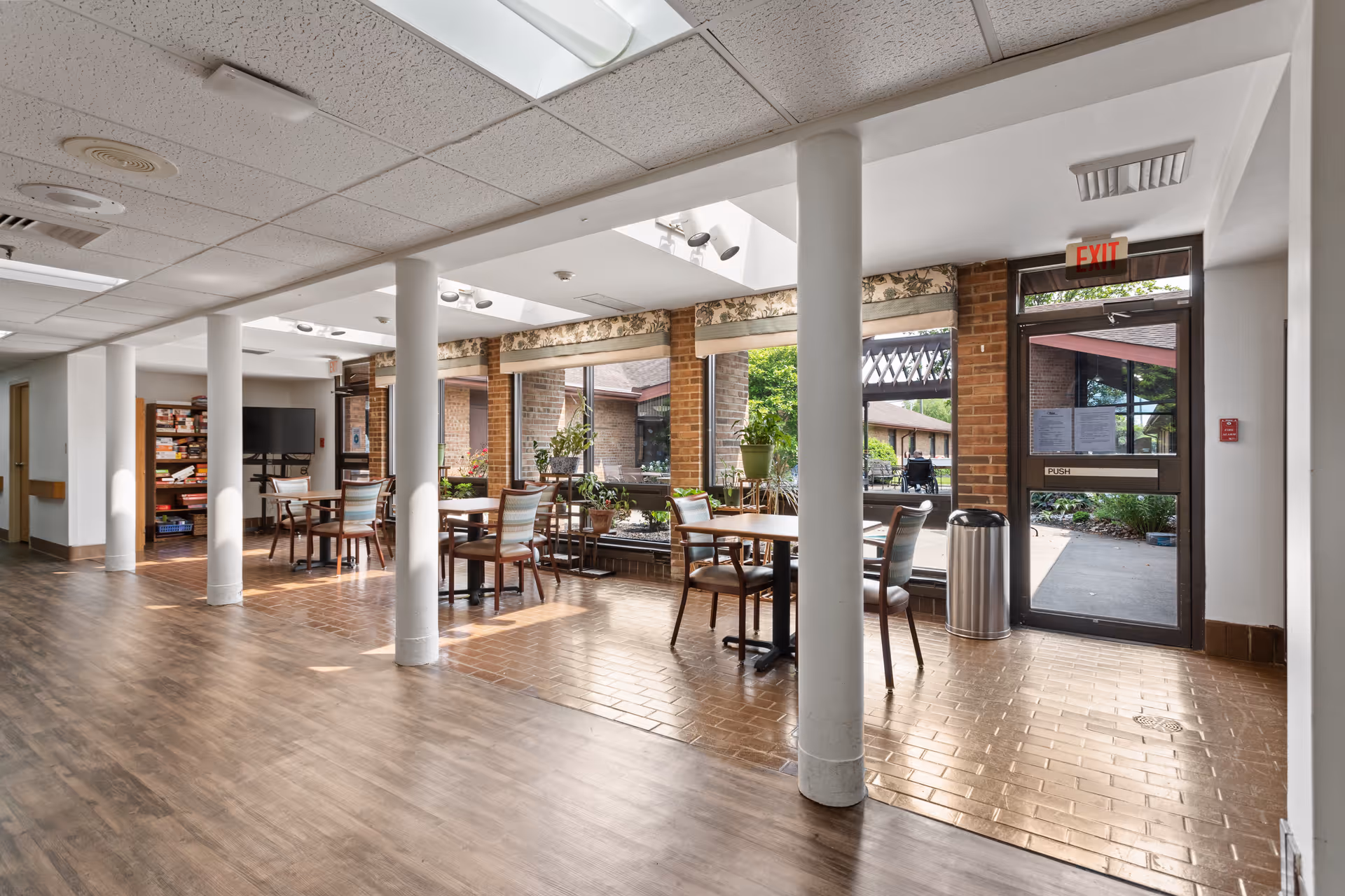 A bright common area in a senior living facility with several tables and chairs arranged near large windows that look out onto a garden courtyard. The floor is a combination of wood and tile, and there are white support columns throughout the space. A door with an exit sign leads outside, and there is a trash can nearby. Shelves with books and a television are visible in the background.