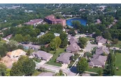 Aerial view of a residential community with multiple single-story buildings surrounded by trees and greenery, with a larger building and a small lake visible in the background.