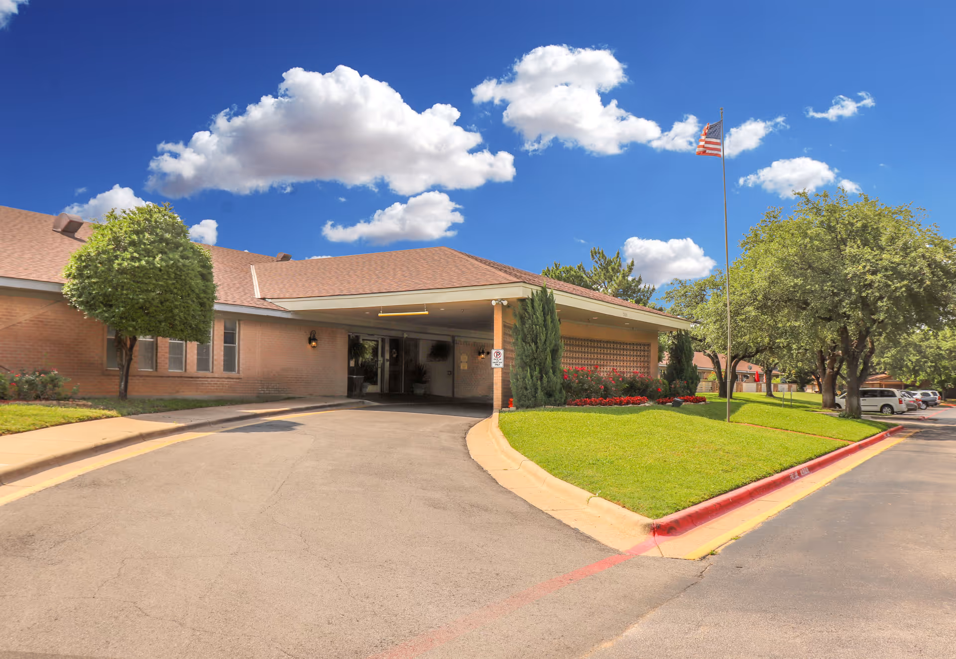 Front entrance of a single-story brick rehabilitation and healthcare facility with a covered drive, flagpole, and landscaped lawn under a blue sky.