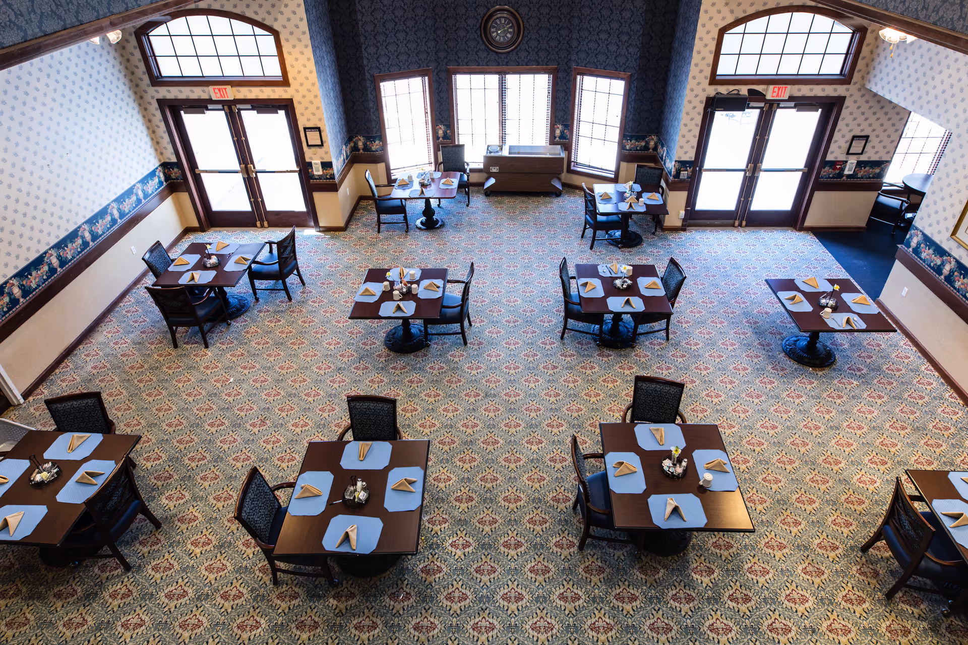 Overhead view of a dining room in an assisted living facility with multiple square tables, each set with blue placemats and folded napkins. The room has patterned carpet, wallpaper with a floral border, large windows, and double glass doors with exit signs above them.