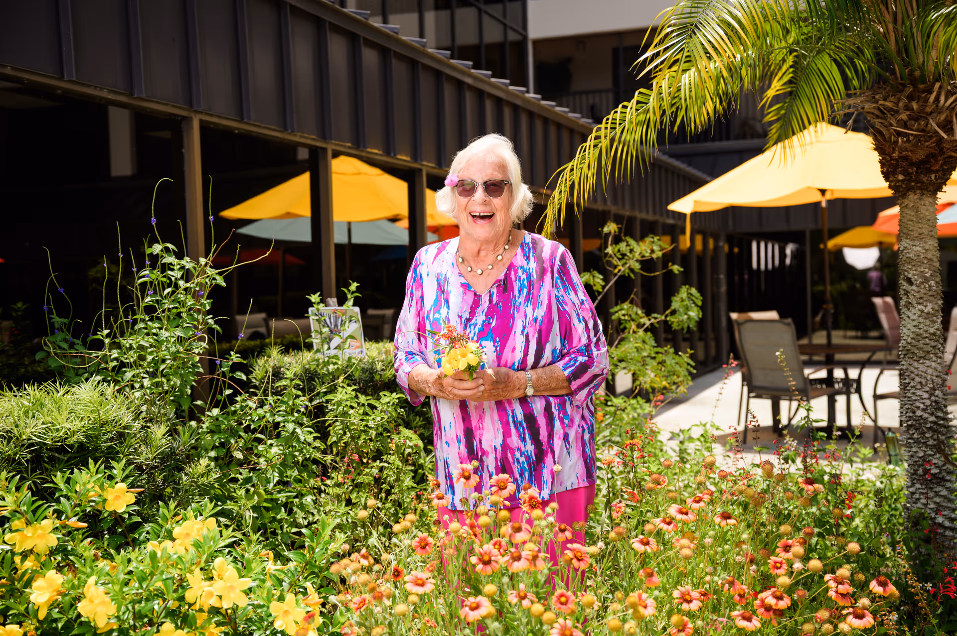 An elderly woman wearing sunglasses and a colorful pink and purple blouse stands smiling in a garden filled with yellow and orange flowers. Behind her are patio tables with yellow umbrellas and a building with large windows.