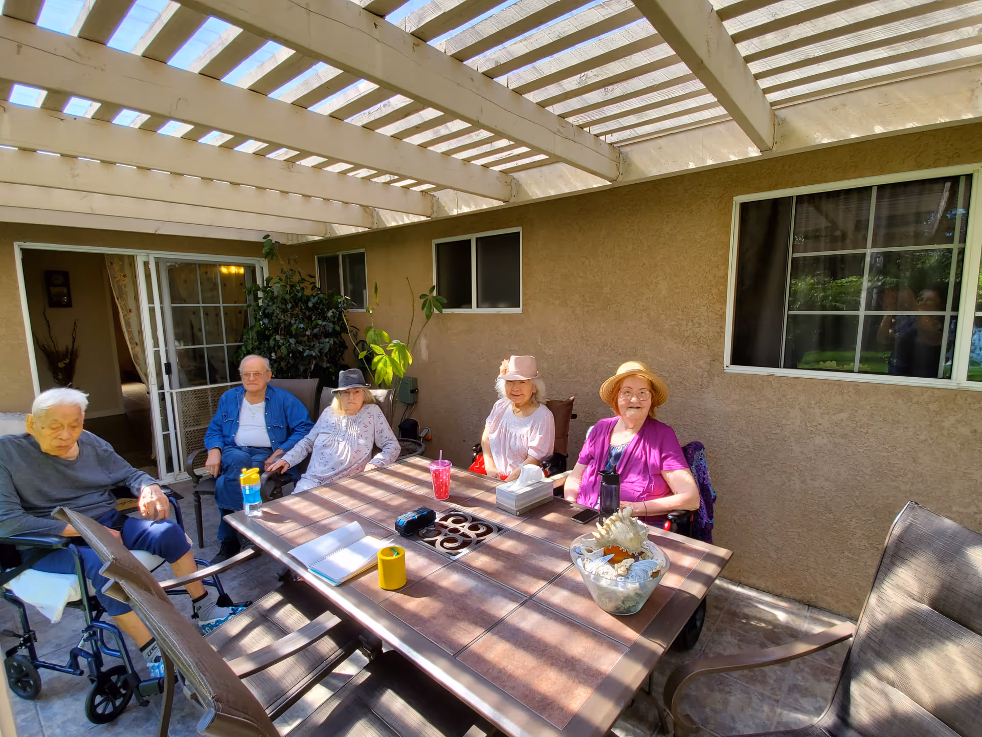 Five elderly people sitting around a rectangular outdoor table under a pergola. The table has various items on it including a notebook, a yellow cup, a pink drink, a tissue box, and a decorative bowl with seashells. The setting is a patio area with beige walls and windows in the background.