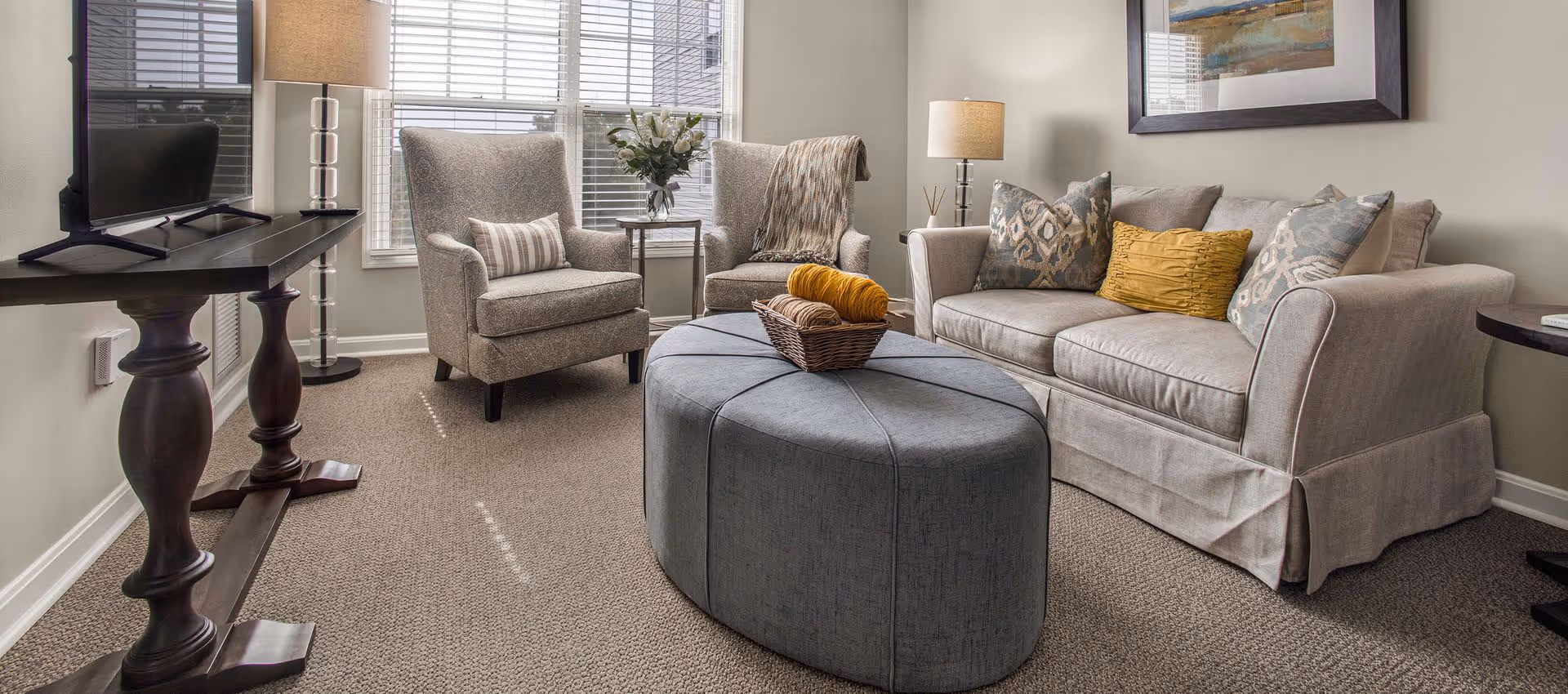 Well-lit living room with a sofa, two armchairs, a round upholstered ottoman, TV on a console, and table lamps.