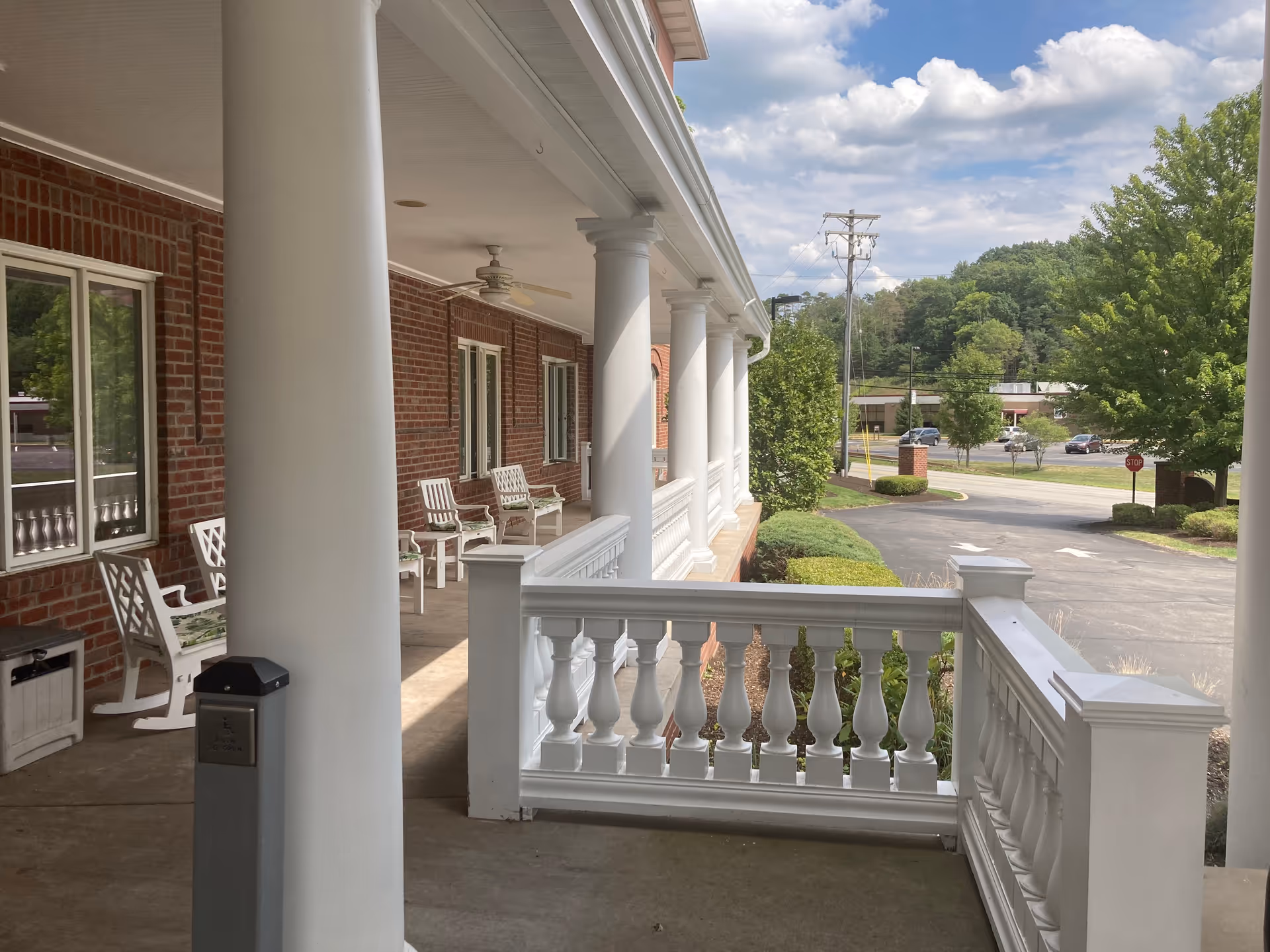 Covered porch area with white columns and white rocking chairs along a red brick building. The porch overlooks a driveway and landscaped greenery with trees and bushes under a partly cloudy sky.