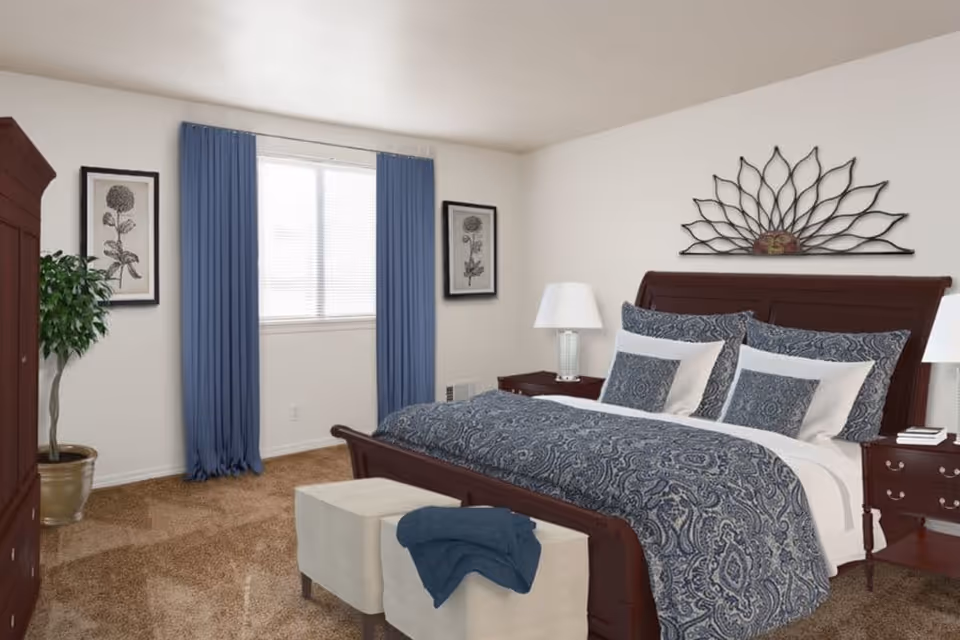 A bedroom with a large wooden bed featuring blue and white patterned bedding and multiple pillows. There are two matching wooden nightstands with lamps on either side of the bed. A decorative metal wall art piece is hung above the headboard. Blue curtains frame a window in the background, and two framed botanical prints hang on the wall. A potted plant is placed in the corner, and a beige ottoman with a blue blanket is at the foot of the bed. The room has beige carpet flooring and white walls.