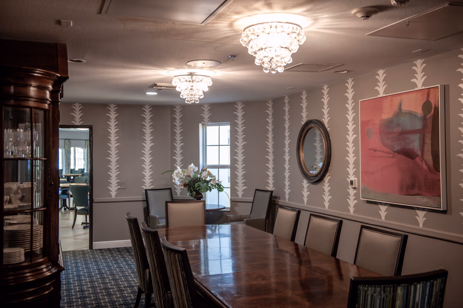 A formal dining room with a long wooden table surrounded by chairs. The walls are decorated with patterned wallpaper, a round mirror, and a large abstract painting. Two chandeliers hang from the ceiling, and a cabinet with glassware and plates is visible on the left side. A window and an open doorway leading to another room are also visible.
