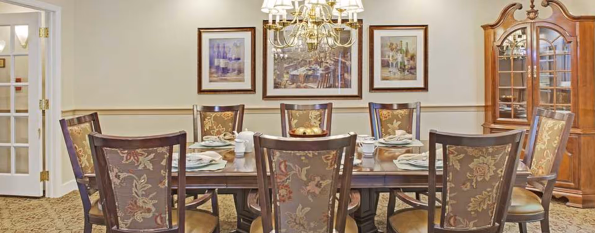 Formal dining room with a large wooden table set for a meal, upholstered chairs, chandelier, framed artwork, and a china cabinet.