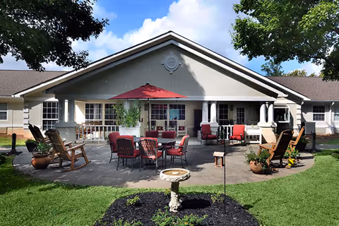 Outdoor patio area of a senior living facility with multiple seating arrangements including red cushioned chairs around a table with a red umbrella, wooden rocking chairs, potted plants, and a birdbath in the foreground. The building has a light-colored exterior with white columns and large windows under a peaked roof.