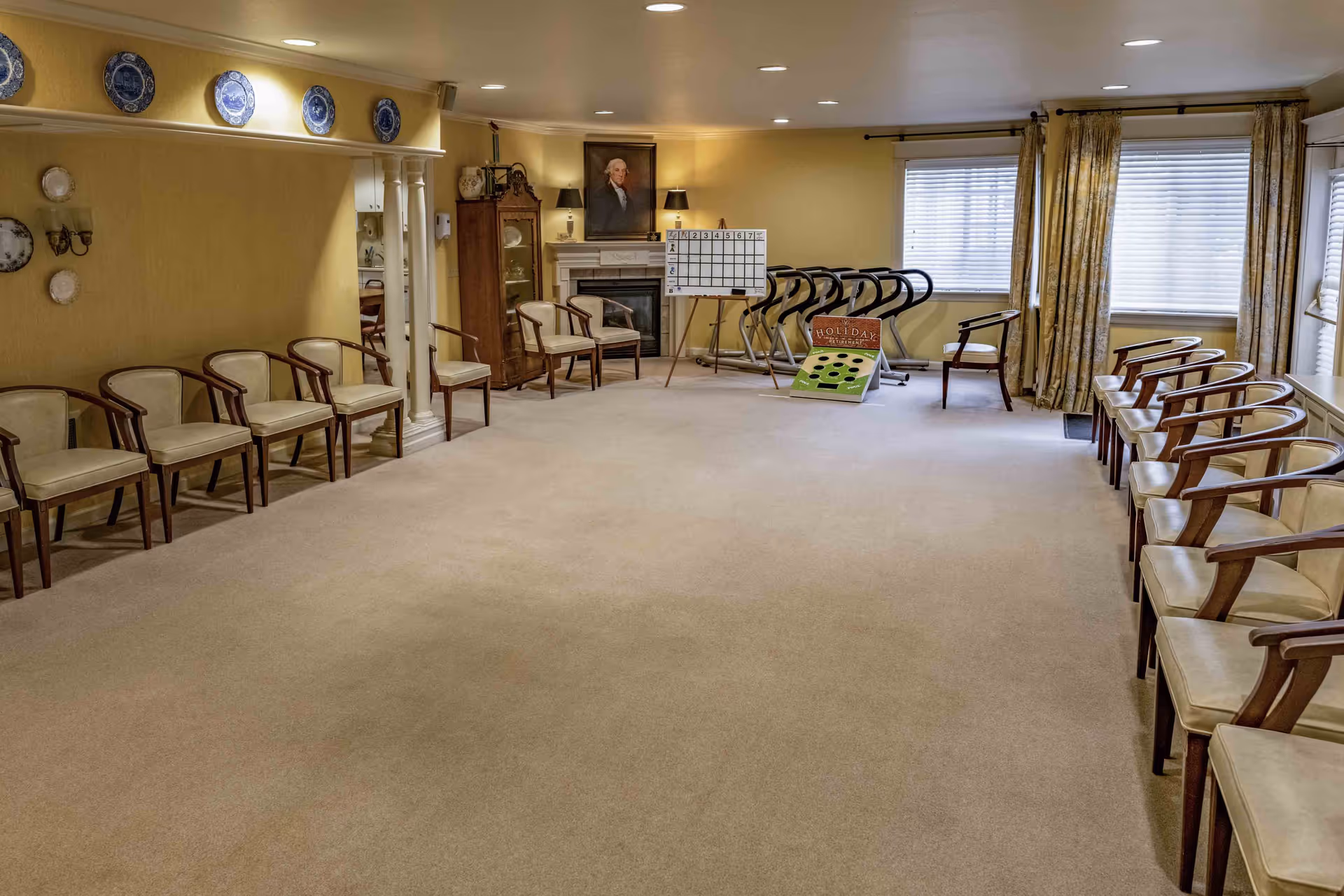 A spacious room with beige carpet and yellow walls, featuring two rows of wooden chairs with cream cushions lined up along the left and right walls. At the far end, there is a fireplace with a portrait above it, a cabinet, and a whiteboard on an easel. Several exercise machines are positioned near a window with closed blinds and floral curtains. The room is softly lit with ceiling lights and wall sconces.