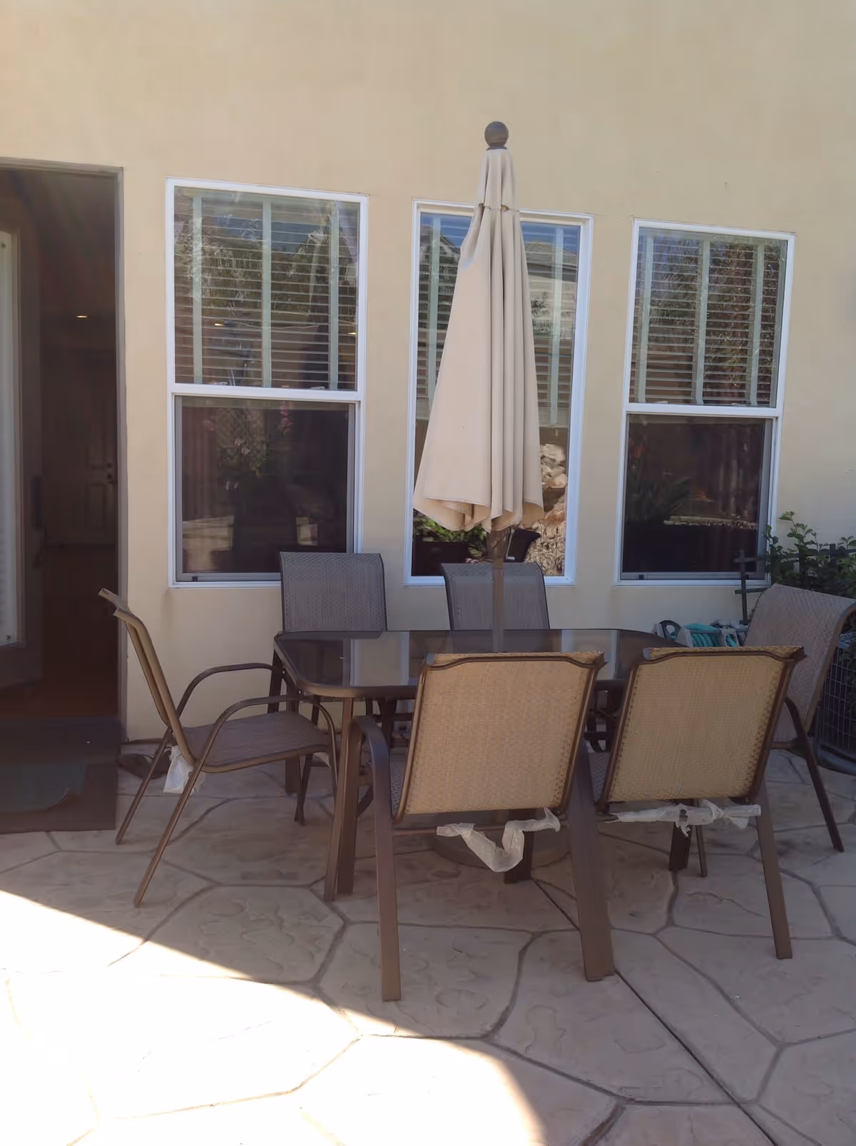 Outdoor patio area with a glass-top table surrounded by six chairs and a closed beige patio umbrella in the center. The patio has a stone tile floor and is adjacent to a beige building with three windows and an open door.