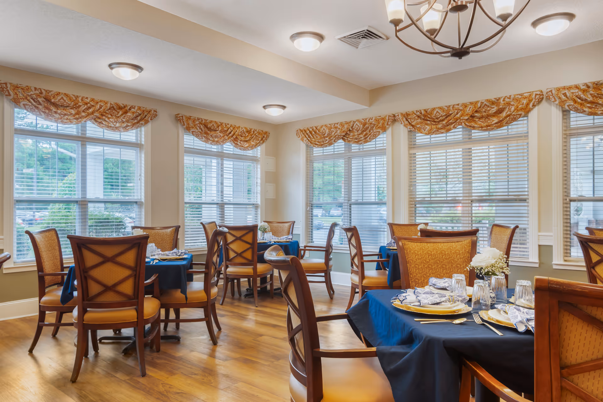 A bright dining room with multiple tables covered in dark blue tablecloths, each set with plates, napkins, glasses, and silverware. The room has large windows with white blinds and patterned valances, wooden chairs with cushioned seats, and a wooden floor. A chandelier and ceiling lights illuminate the space.
