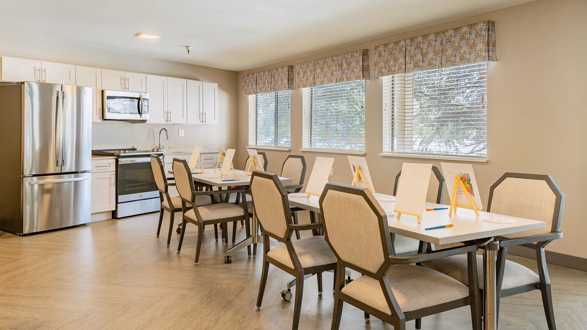 Well-lit communal dining/activity room with tables and chairs set up for painting and a small kitchen with stainless steel appliances.