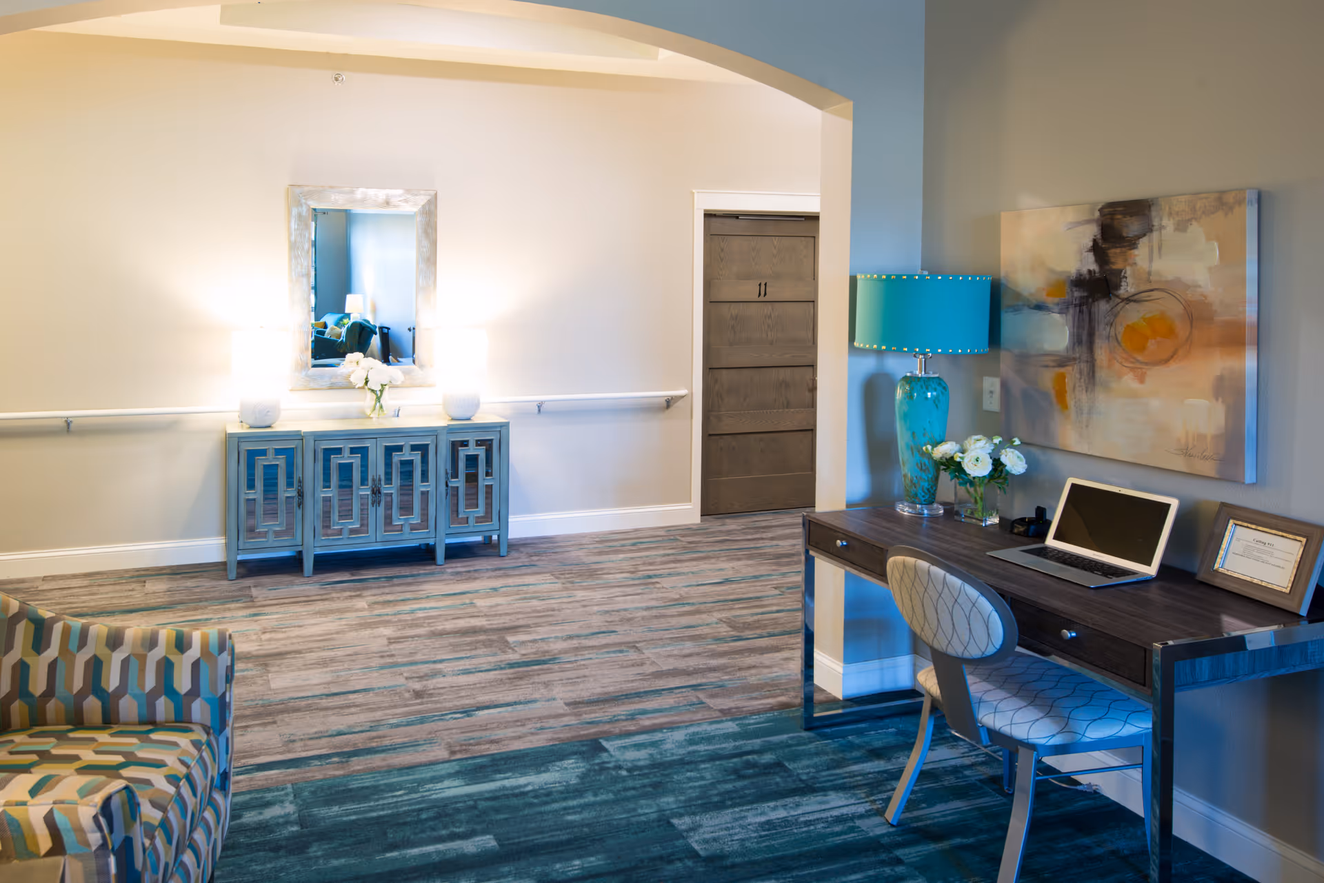 A well-lit interior room featuring a patterned armchair on the left, a blue decorative cabinet with a mirror above it on the back wall, and a wooden desk with a laptop, a turquoise lamp, flowers, and a framed certificate on the right. The floor has a mix of wood and carpeted sections, and there is a closed wooden door in the background.