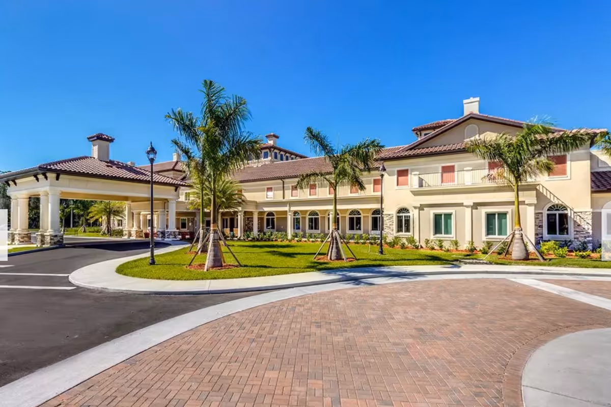 Exterior view of a large, two-story senior living facility building with beige walls and a red-tiled roof, surrounded by palm trees and a well-maintained lawn under a clear blue sky. There is a circular driveway with brick paving in front of the entrance.