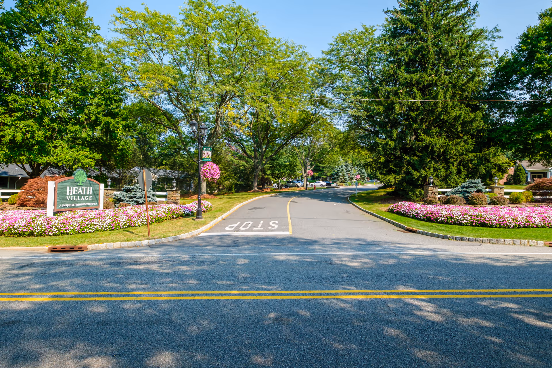 Entrance to Heath Village Retirement Community showing the driveway, landscaped flowerbeds, trees, and a 'Heath Village' sign.