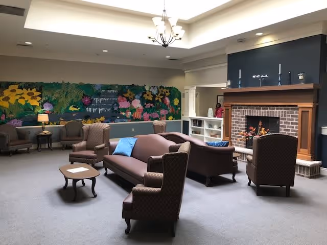 A spacious living room area with multiple upholstered chairs and sofas arranged around a wooden coffee table. The room features a brick fireplace with a wooden mantle decorated with candles and small items. A colorful mural of flowers and greenery adorns the far wall. The ceiling has recessed lighting and a chandelier, and there is a small side table with a lamp near the mural.