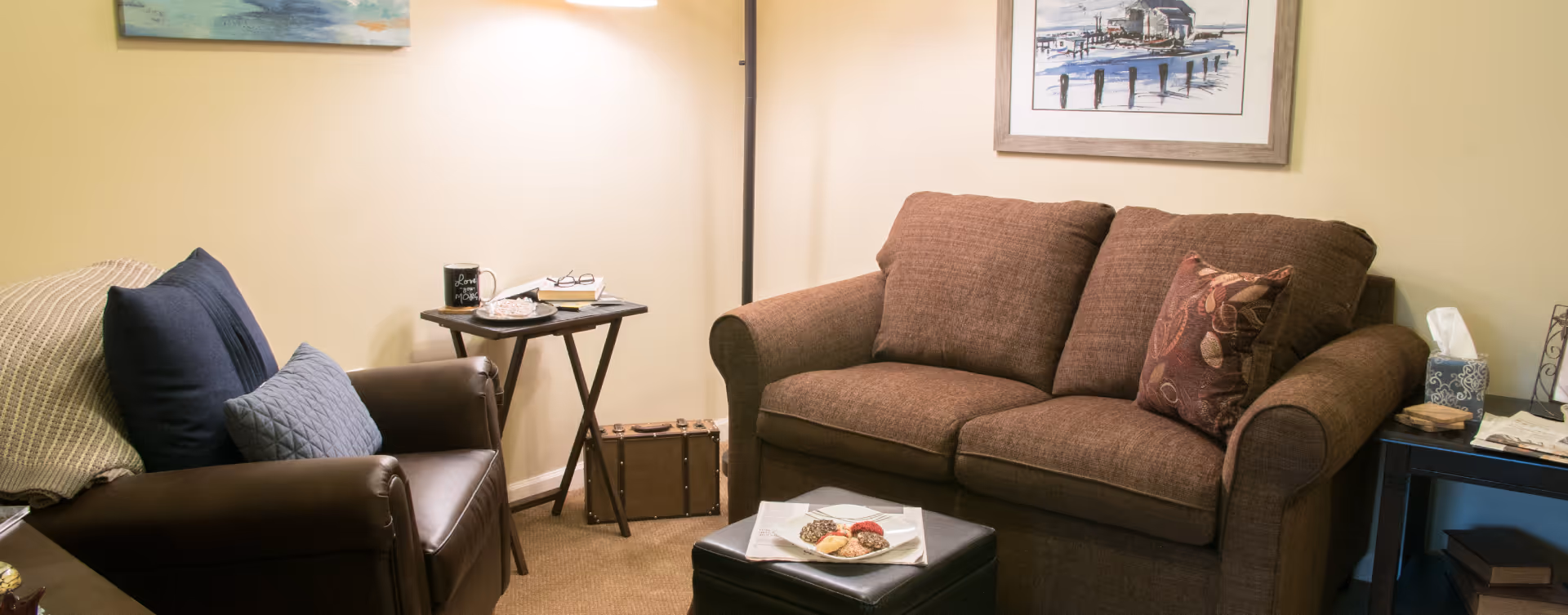 A cozy living room with a brown fabric sofa adorned with a decorative pillow, a dark brown leather armchair with blue cushions, a small side table holding a mug, glasses, and a book, a floor lamp, and framed artwork on the beige walls. A black ottoman with a plate of cookies is in front of the sofa.