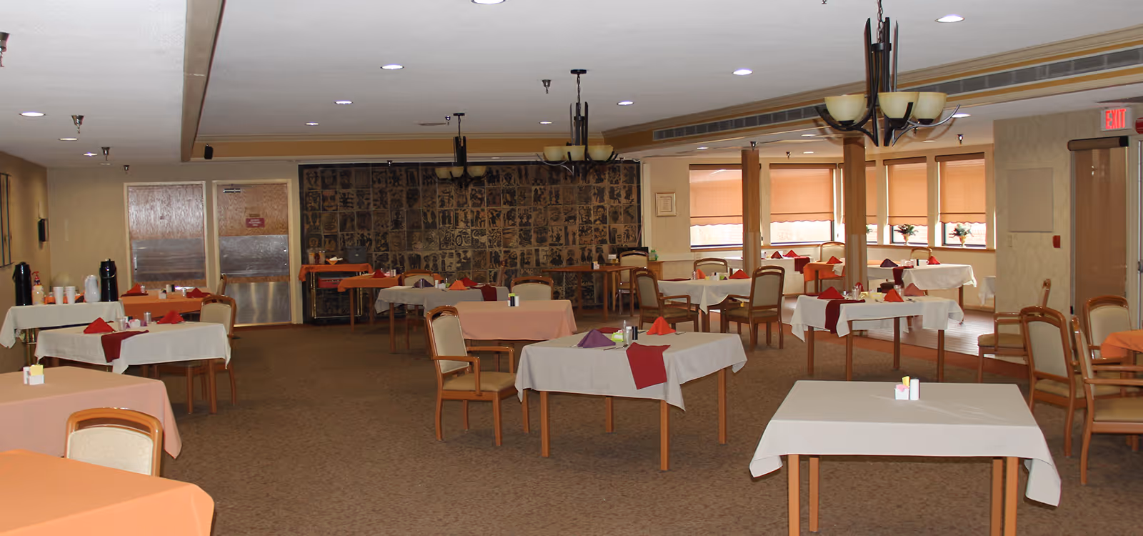 A spacious dining room with multiple tables covered in white and peach tablecloths, each set with folded napkins in red and purple. Wooden chairs with cushioned seats surround the tables. The room features beige walls, a carpeted floor, large windows with blinds, and ceiling light fixtures. There is a metal door and a beverage station with coffee urns on the left side.