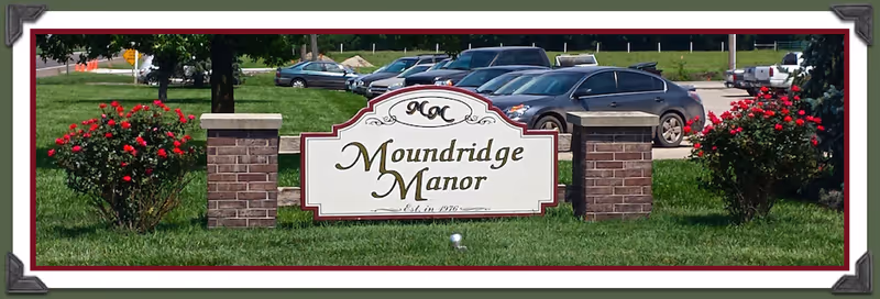 A white and maroon sign with the text 'Moundridge Manor Est. in 1976' set on a green lawn with two brick pillars on either side. Behind the sign, there is a parking lot with several parked cars and some trees in the background.