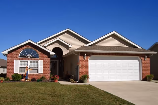 Single-story residential house with a brick and beige siding exterior, a large white garage door, a front entrance with an arched doorway, and a well-maintained lawn under a clear blue sky.