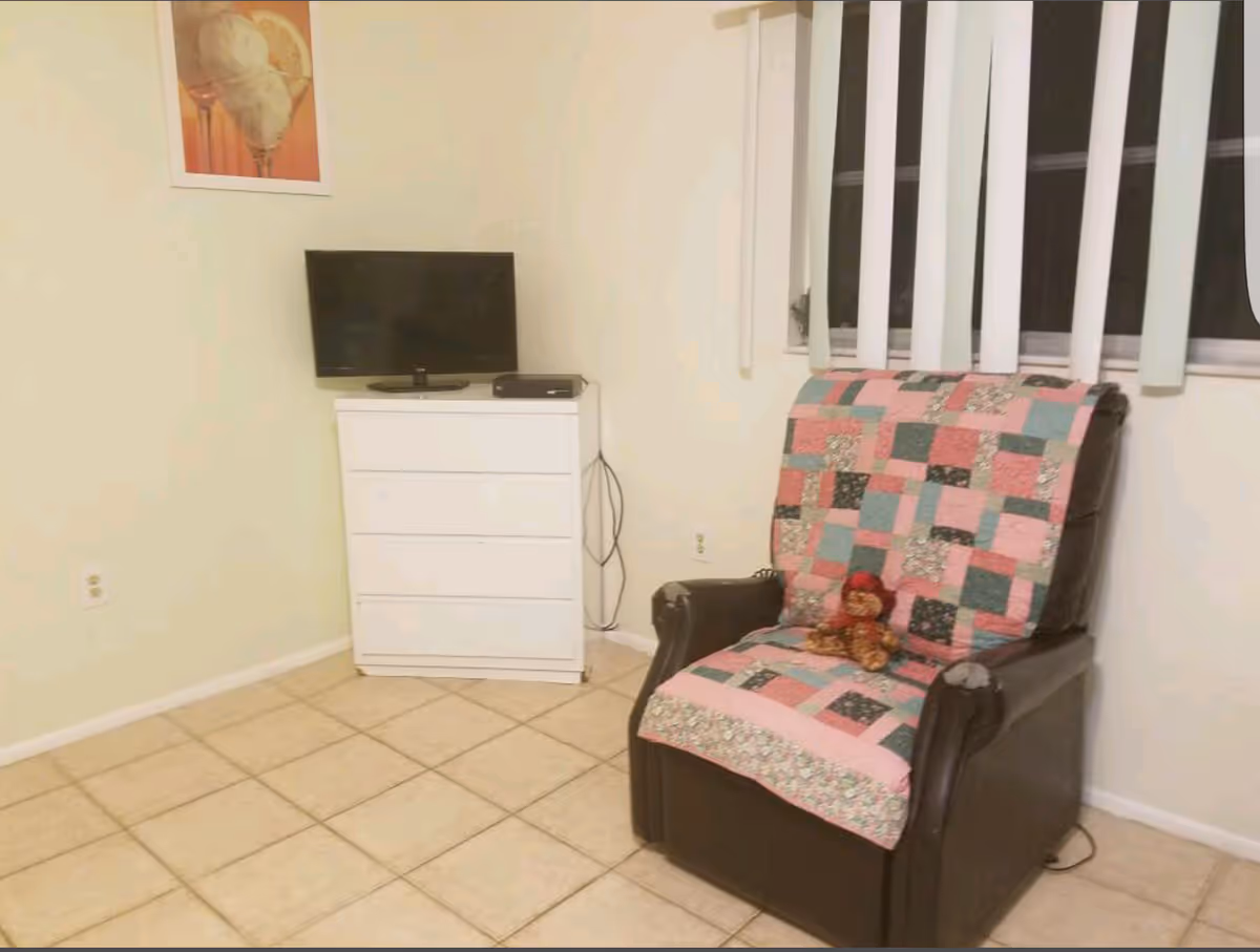 A small living area with a TV on a white dresser and a dark recliner covered by a pink and green patchwork quilt and a teddy bear, next to a window with vertical blinds.