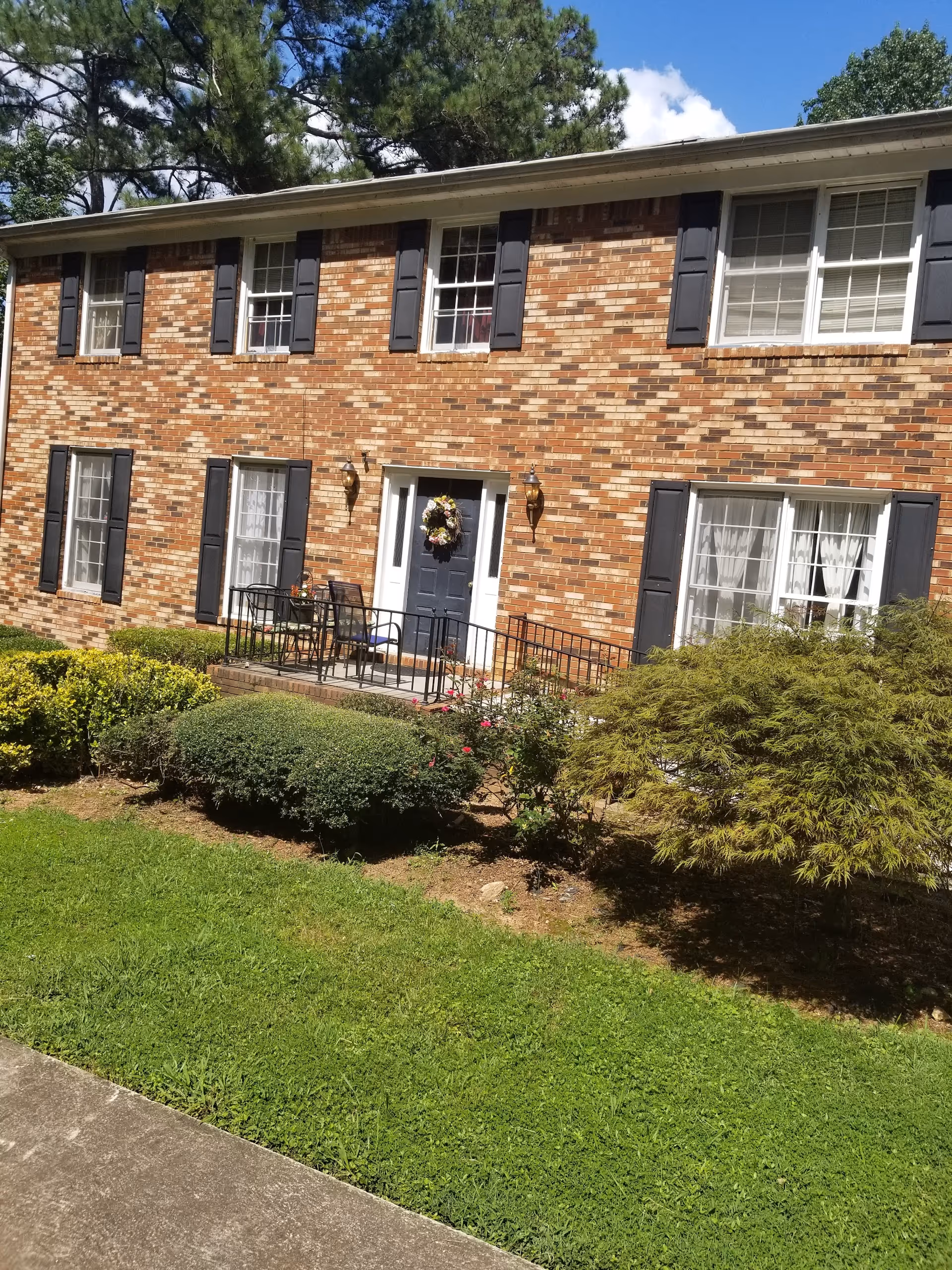 Two-story brick house front with black shutters, a central door with a wreath, small porch and trimmed lawn and shrubs.