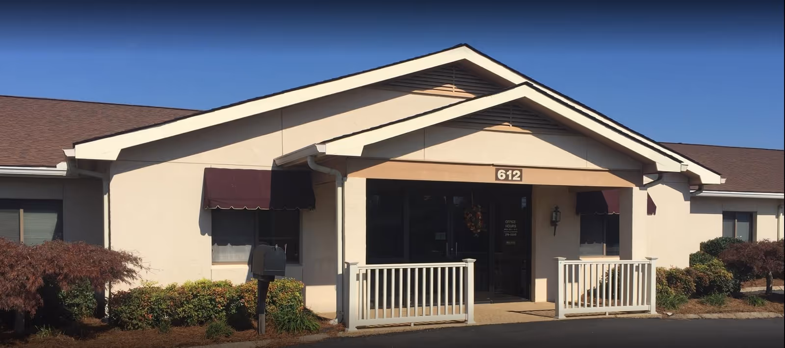 Exterior front view of a single-story building with beige walls and a brown roof. The entrance has a small porch with white railings and a sign above the door displaying the number 612. There are two windows with maroon awnings on either side of the entrance, and landscaping with bushes and small trees in front of the building. The sky is clear and blue.