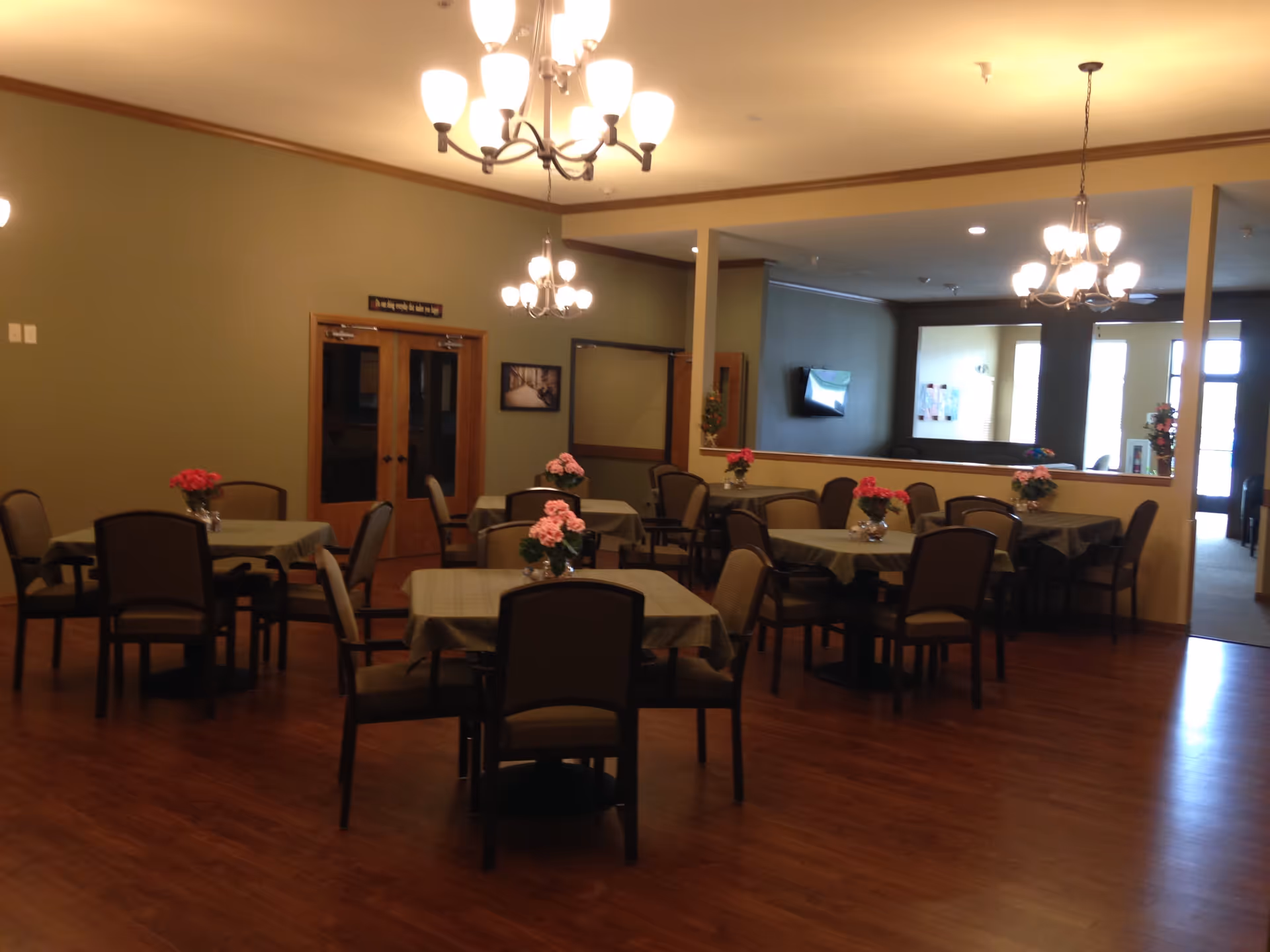 A dining room with several square tables covered with green tablecloths, each table decorated with a vase of pink flowers. The room has wooden floors, beige walls, and multiple chandeliers providing warm lighting. There are wooden double doors and a large mirror on one wall reflecting part of the room and a TV mounted on the opposite wall.
