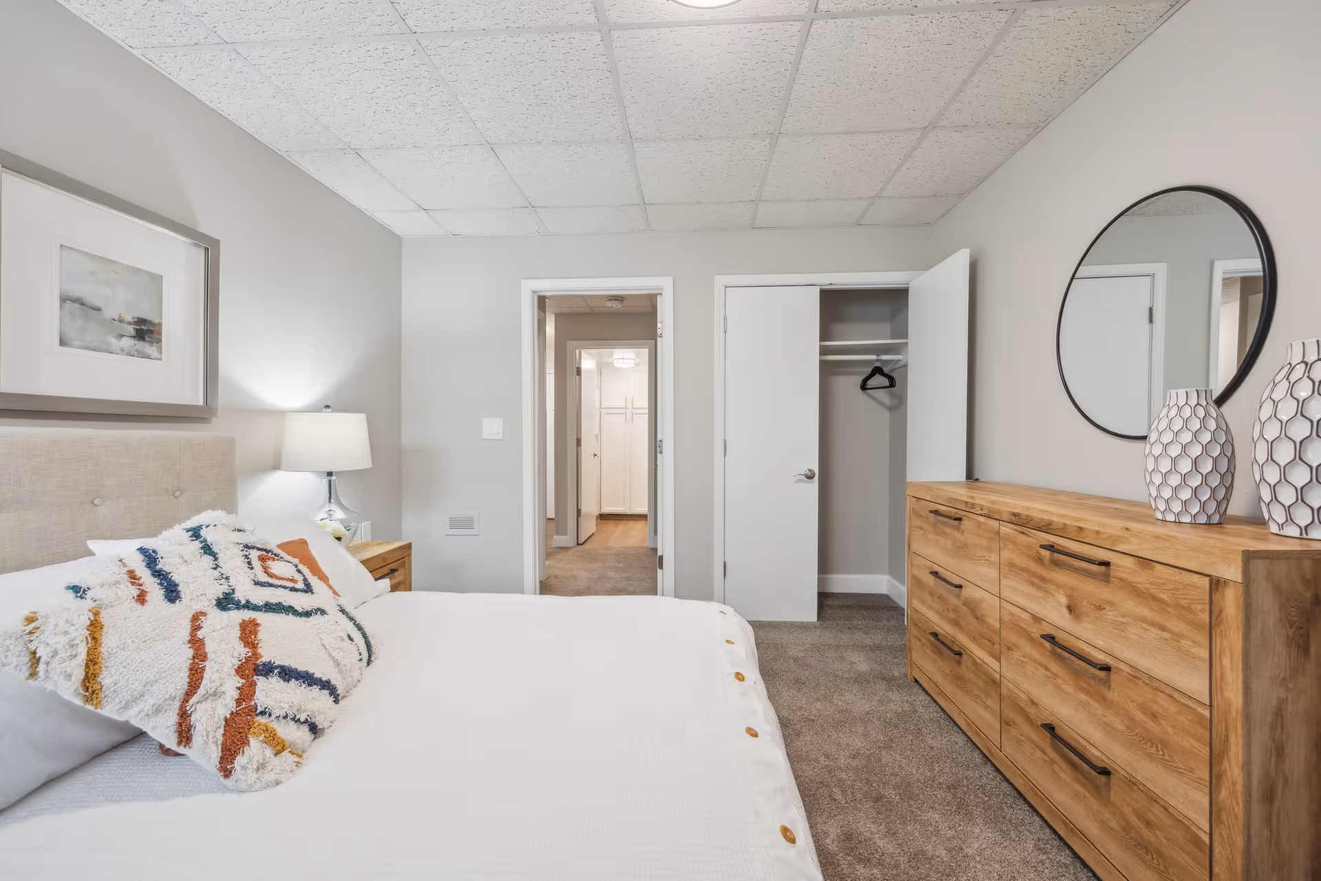 A neatly arranged bedroom with a bed featuring a white comforter and a colorful decorative pillow. There is a wooden nightstand with a lamp on the left side of the bed. Opposite the bed is a wooden dresser with six drawers, decorated with two patterned vases and a round mirror above it. The room has light gray walls, a carpeted floor, and a ceiling with white tiles. An open closet door reveals a single black hanger inside, and a doorway leads to a hallway with additional doors visible.