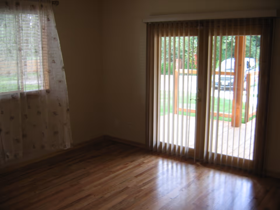 Empty room with wooden floor, a window with sheer curtains on the left, and glass sliding doors with vertical blinds leading to an outdoor deck area.