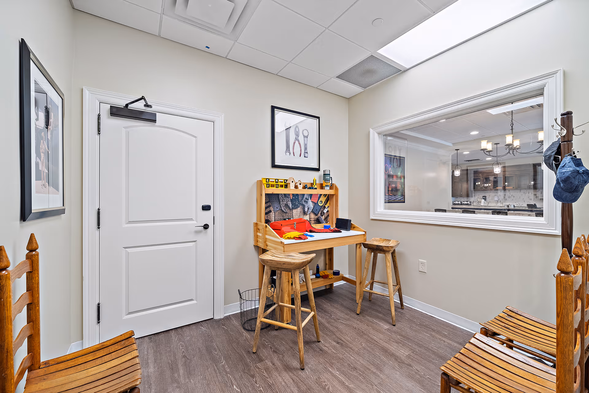 A small room with light-colored walls and wood flooring featuring a children's play workbench with toy tools, two wooden stools, and four wooden chairs. There is a large window looking into an adjacent room with a chandelier and kitchen area visible. A white door is on the left wall, and a coat rack with hats is on the right side.