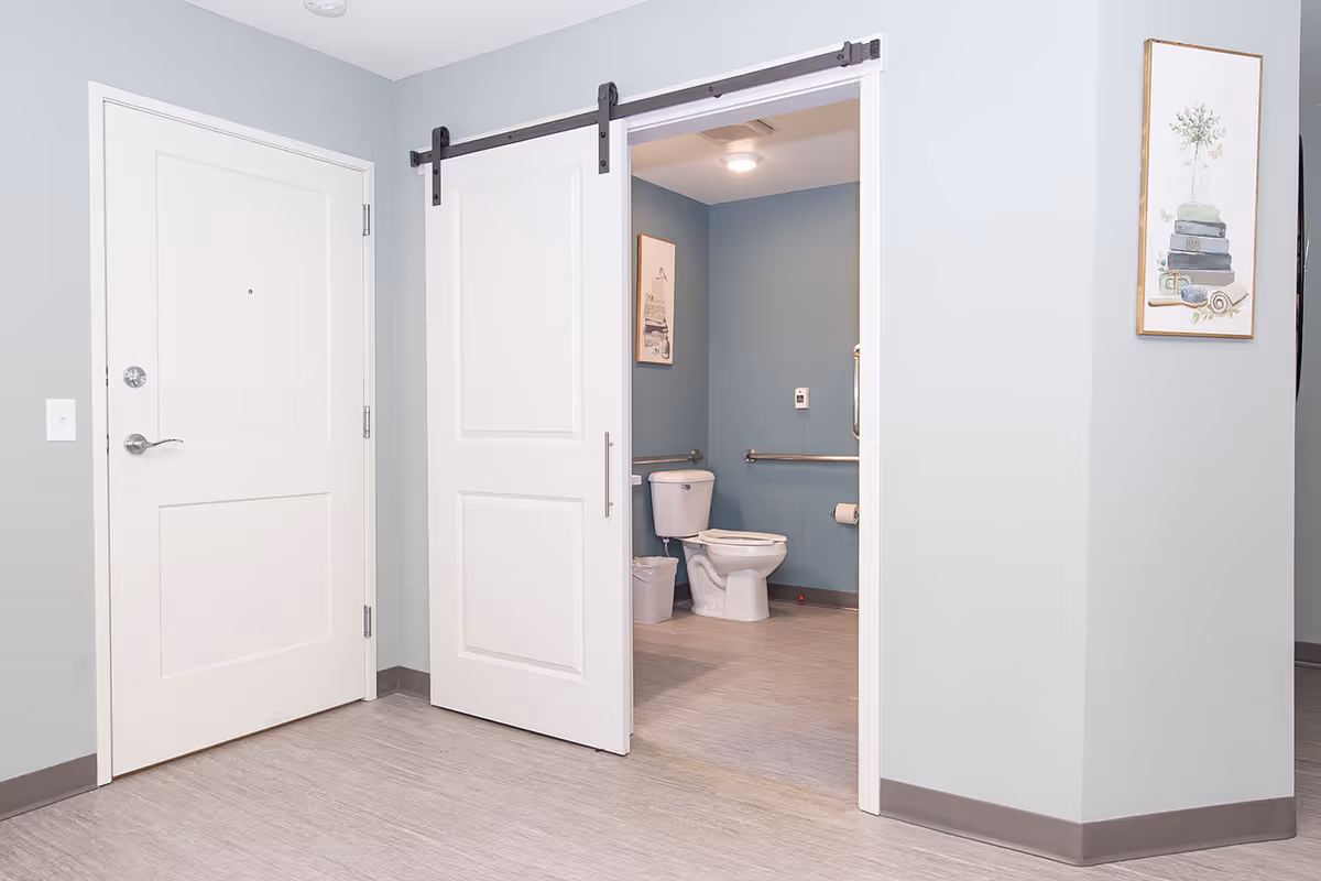 Interior view of a room with light gray walls and flooring, featuring a white sliding barn door partially open to reveal a bathroom with a toilet, grab bars, a trash can, and a framed picture on the blue wall.
