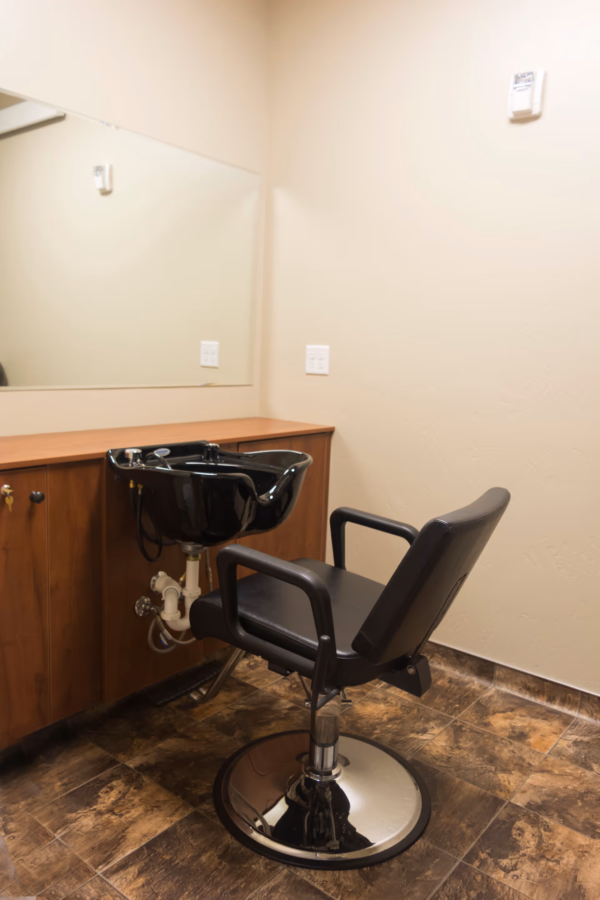 Interior view of a hair washing station with a black salon chair and a black sink basin attached to a wooden cabinet. A large mirror is mounted on the wall above the cabinet, and the floor has brown patterned tiles.