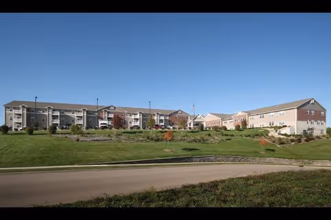 Wide exterior view of Oak Park Place Dubuque senior living facility showing multiple connected buildings with beige and brick facades, surrounded by green lawns and small trees under a clear blue sky.