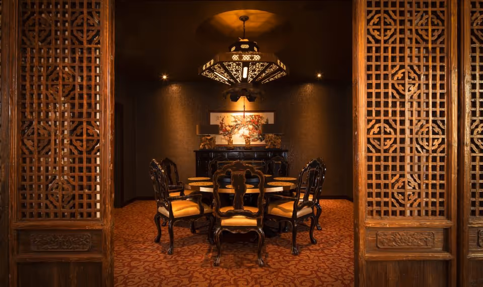 Ornate Asian-style dining room with a round table and carved wooden chairs framed by lattice wood screens and a decorative hanging light.