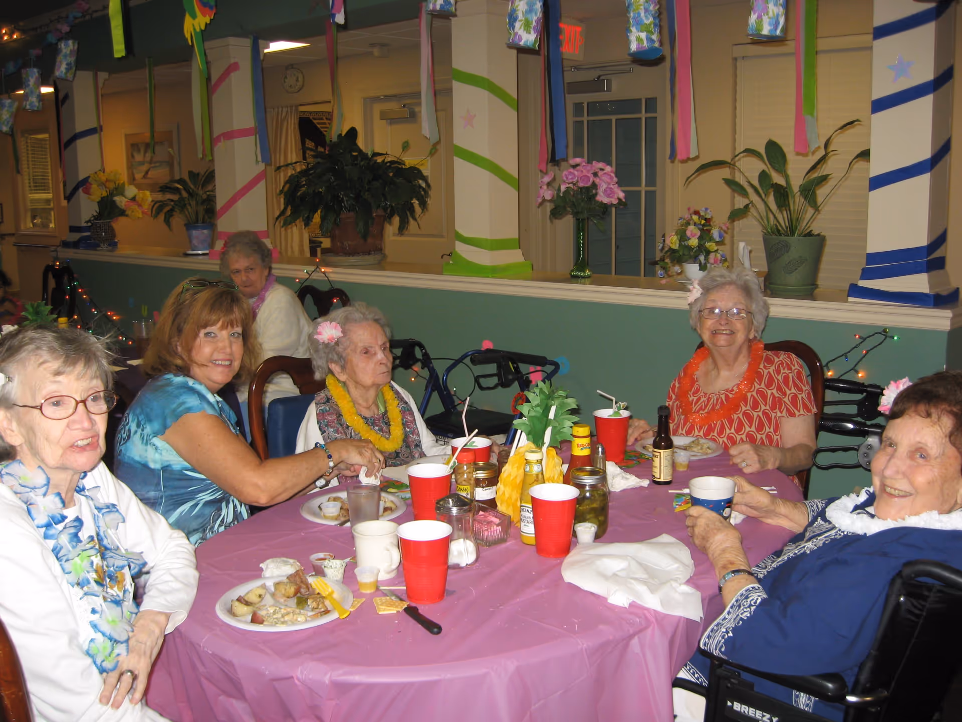 A group of elderly women and a caregiver sitting around a table covered with a pink tablecloth, enjoying a meal together in a decorated room with colorful streamers and plants. Some women are wearing leis and flowers in their hair, and there are various condiments and drinks on the table.