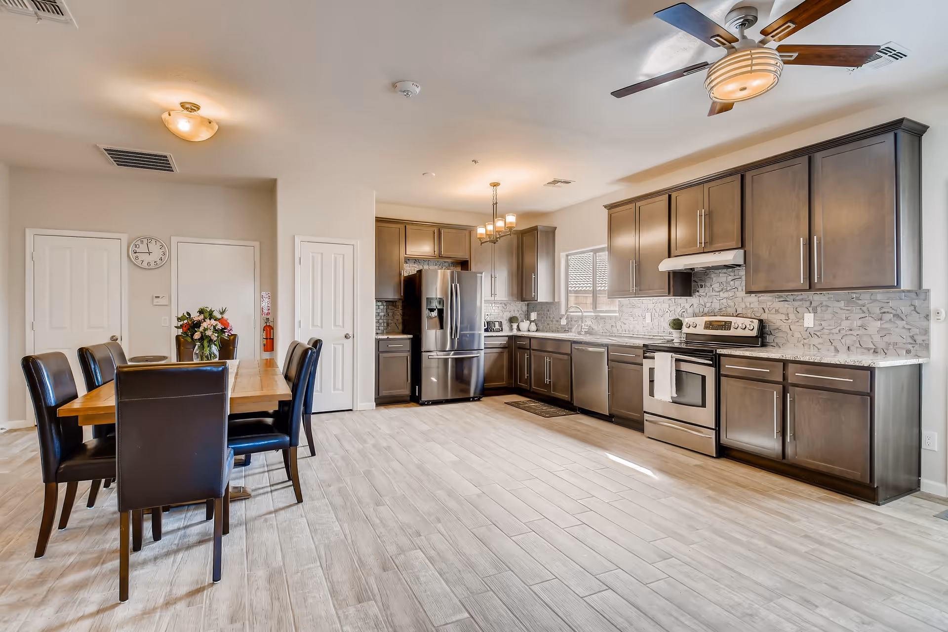 A spacious and modern kitchen and dining area in an assisted living facility. The kitchen features dark wood cabinets, stainless steel appliances including a refrigerator, stove, and dishwasher, and a tiled backsplash. The dining area has a wooden table with six black leather chairs and a vase of flowers on the table. The room has light-colored tile flooring, a ceiling fan, and multiple light fixtures.